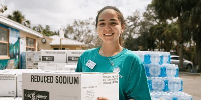 In front of a building, a woman carries boxes of emergency food for an outdoor food distribution.