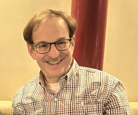 Lester Nathan, smiling with glasses and a checkered shirt, seated with a red pillar in the background.